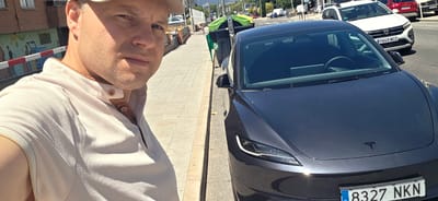 In the city of Collado Villalba, Spain, a man wearing a light-colored collared shirt and a baseball cap takes a selfie. He is standing on a sidewalk next to a dark-colored Tesla electric vehicle. The car is parked on the street, indicated by the presence of other vehicles and trash bins visible in the background. The sidewalk is made of concrete pavers and is lined with a low wall. Buildings are visible on the left side of the image, and a street with traffic is on the right. The scene appears to be a typical urban environment on a sunny day.