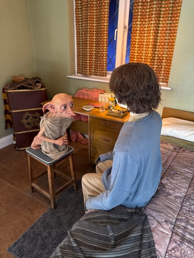 The media file shows an indoor scene, possibly a bedroom or living space. A mannequin with dark, curly hair is partially visible, wearing a light blue crewneck sweatshirt and tan pants. To the right of the mannequin, a bed with patterned bedding is present. In the foreground, a figure resembling Dobby, a character from the Harry Potter series, sits on a wooden chair with a blue cushion. Dobby is wearing a grey, tattered garment. Behind Dobby, a dark red trunk with gold trim and the Hogwarts crest is visible, along with a striped scarf. A wooden dresser with two drawers is situated behind Dobby and to the left. On top of the dresser are several items, including a glass jar with a yellow lid, a small dish, and other indistinct objects. A window with plaid curtains in shades of brown and orange is to the left of the frame. The overall impression is a staged scene, likely a fan-made exhibit or a themed room. The lighting suggests it is daytime. There is no explicit indication of the weather. The presence of the Hogwarts crest and Dobby suggests a Harry Potter theme.