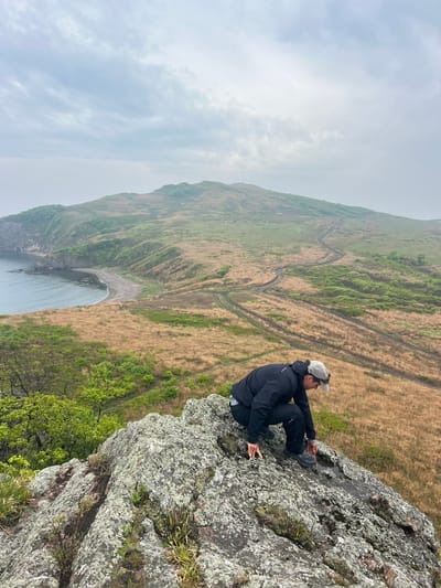 The image shows a man sitting on a rocky cliff overlooking a landscape. The man is wearing a dark jacket, dark pants, a light-colored baseball cap, and dark shoes. He is sitting cross-legged and looking out at the view. The cliff is made of grey rock and is covered in some moss and small plants.

In the background, there is a hillside with a mixture of green and brown vegetation. A winding dirt track can be seen traversing the hillside. At the bottom of the hill is a small beach and the ocean, which stretches out to the horizon. The sky is overcast.

The location is identified as being in the city of Gvozdevo, Russia.