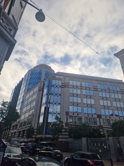 The image depicts a street scene in the municipality of Saint-Gilles - Sint-Gillis, Belgium, dominated by a large, modern building with a facade of light-colored stone and numerous reflective blue windows. The building appears to be an office or commercial structure, with several stories visible. In the foreground, traffic is present, with several cars, including a dark-colored hatchback with a visible license plate starting with "1 XAS," and a motorcycle. A pedestrian crossing signal and parking signage are also visible. Trees and foliage line the street in front of the building, and a metal fence encloses a portion of the property. Overhead, a street lamp and power lines are visible against a cloudy sky. The overall impression is that of a busy urban environment.