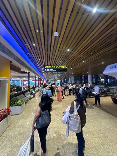 The image depicts an indoor airport terminal concourse, identified as being in Kasarvarne, India. The environment features a high ceiling constructed with light-colored, horizontal wooden slats, interspersed with recessed circular lights and a linear blue LED accent light strip along the upper left wall. The floor is a polished light-colored tiled surface.

Approximately 25-35 individuals are present within the concourse. In the foreground, two women walk away from the camera. The woman on the left has long dark hair, wears dark clothing, and carries a grey shoulder bag and a white bag in her left hand. The woman on the right has dark hair tied back, wears a brown top, and carries a light-colored backpack with a light jacket draped over it.

In the mid-ground, a diverse crowd of people occupies the concourse, moving in various directions. Attire includes a mix of casual clothing, such as a person in an orange-pink garment and another in a straw hat, and semi-formal wear. Many individuals are carrying personal bags, including backpacks and shoulder bags.

Overhead, a black rectangular sign with yellow text indicates "Gates / द्वार 11-13, 21-23" with a right-pointing arrow, and "Toilets / शौचालय / ಶೌಚಾಲಯ" with a left-pointing arrow.

On the left side of the concourse, several silver-colored public seating benches are visible, some occupied. Planters containing green foliage and red flowers are positioned along this edge. Beyond these, commercial storefronts or lounge areas with interior lighting are visible. On the right side, further into the background, rows of unoccupied or sparsely occupied airport seating are present alongside several pieces of luggage. A man in a white shirt and dark trousers stands near these seats.