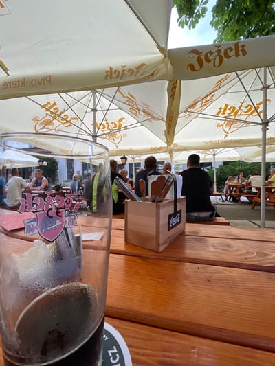 The image depicts an outdoor seating area of a beer garden, likely located in Jihlava, Czechia, under large white umbrellas that bear branding. In the foreground, a glass of dark beer with foam sits on a wooden table, partially obscuring a colorful sticker on the glass with a hedgehog logo and the text "Jezek 18". A wooden holder containing cutlery and a Coca-Cola logo is also on the table. Several patrons are seated at picnic tables in the background, enjoying drinks and socializing. The environment suggests a relaxed, casual atmosphere, possibly during daytime or early evening.