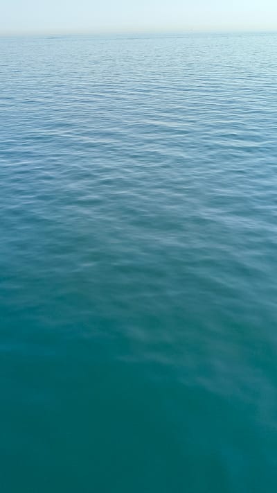 The image shows a vast expanse of calm, blue-green water stretching towards a hazy horizon. The surface of the water is gently rippled, creating a textured pattern that shifts from a lighter blue near the horizon to a deeper teal in the foreground. The sky is a pale, almost white hue, with a faint suggestion of distant land or structures on the far horizon. There are no people, animals, or distinct objects visible in the scene. The overall impression is one of tranquility and open space. The location in Unknown, France is not specified in the image.
