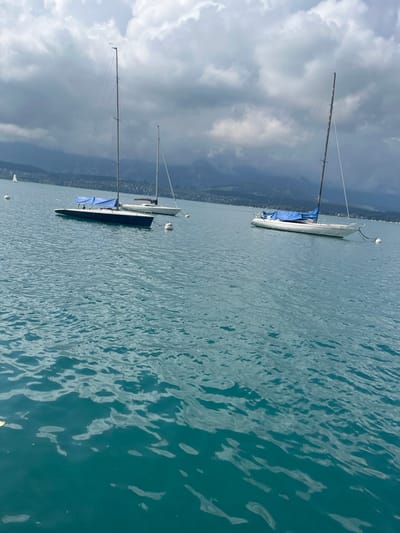 The image displays a segment of Lake Thun in Hünibach, Switzerland, under an overcast sky with prominent grey cloud formations. The water in the foreground and mid-ground is a teal-green color, exhibiting a rippled surface. Three sailboats are moored. The most prominent vessel, located in the left-midground, features a dark-colored hull, a tall mast, and a blue sail cover secured over its boom. A second white-hulled sailboat, also equipped with a mast and a sail cover, is moored slightly behind and to the right of the dark-hulled boat. Further right, a third white-hulled sailboat, with a mast and a blue sail cover, is similarly moored. Multiple white cylindrical mooring buoys are visible in the water, positioning the vessels. The background reveals a distant shoreline with green vegetation and structures, backed by mountainous terrain, partially obscured by the low cloud cover. A smaller white sailboat is observed near the distant left shore.