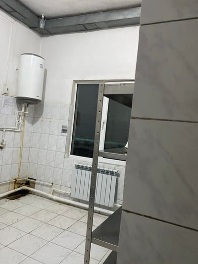 The image depicts an interior view of what appears to be a utility room or bathroom, likely in Ranchpar, Armenia. The room is predominantly tiled with white square tiles on the walls and floor. A white cylindrical water heater, branded "BAXI", is mounted on the wall, with visible plumbing connections.
A window with dark tinted glass is set into one wall, reflecting some of the interior. Adjacent to the window, a white radiator with horizontal fins is visible, suggesting a heating system. A metal railing or shelf with evenly spaced holes runs horizontally across the image, appearing to be attached to the wall near the window.
On the left side of the image, a large grey rectangular duct runs vertically, indicating ventilation or an exhaust system. There are various pipes and conduits visible throughout the space, suggesting functional utility.
There is no activity or event taking place, as the scene is static and appears unoccupied. The lighting is uniform and bright, typical of an indoor electric light source, making it difficult to determine the time of day. No specific weather conditions are discernible.
A small piece of paper with text and an image is affixed to the tiled wall below the window, but the text is illegible. Another document or label is partially visible near the base of the water heater. The overall impression is one of a functional, somewhat utilitarian space.