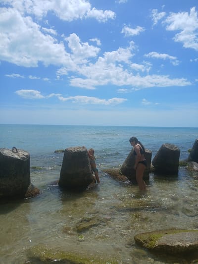 The image depicts two individuals interacting with a body of water, likely the Black Sea given the location context. The scene is a coastal environment with large concrete blocks serving as breakwaters or a pier. The sky is a clear blue with scattered white cumulus clouds, suggesting fair weather. The water is clear and shallow around the concrete structures, with sunlight reflecting off its surface.

One person, appearing to be an adult female, is positioned on one of the concrete blocks, wearing a black bikini. She is leaning forward, looking down at the water. Another individual, possibly a child, is also on a concrete block, lying on their stomach and wearing light-colored shorts. Both individuals appear to be enjoying their time at the seaside. The concrete blocks are rough and weathered, with some patches of green algae or moss visible, indicating they have been in the water for some time. The background shows a vast expanse of the sea meeting the horizon, with the sky above. There is no visible text in the image. The overall impression is a serene summer day at the beach, with the people engaging in casual recreation by the water.