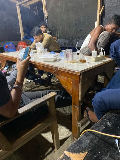 The image captures a casual gathering of several individuals, primarily men, around a wooden table in what appears to be a dimly lit, informal indoor or sheltered setting, likely at night. The scene features a mix of socialization and individual engagement with mobile devices.
Around the wooden table, several objects are visible, including clear plastic cups with straws, some containing beverages, and transparent plastic bags possibly holding food items. A white power strip with multiple charging blocks and cables indicates active device charging. White plastic containers are also present on the tabletop.
At least five people are gathered. One man, wearing a white tank top and light-colored shorts, is seated with his back against a dark wall, looking towards the table. Another individual in blue jeans and a dark top is visible, with their foot, adorned with a black flip-flop, resting on the ground. Towards the left side of the table, a man in a plaid shirt is seen looking down, while another man is partially visible behind him. In the immediate foreground, a person wearing a black top and a silver chain bracelet is engrossed in a smartphone, with another phone visible nearby.
The ground beneath the table appears to be uneven, possibly dirt or rough concrete. Wooden beams are part of the surrounding structure, suggesting a simple or makeshift shelter. A light cloud of smoke drifts in the background on the left, hinting at a nearby cooking fire or heat source. The atmosphere is relaxed, with people appearing to be unwinding and interacting with their phones. Faint text "CE" and "20W" are visible on one of the white charging blocks on the table.