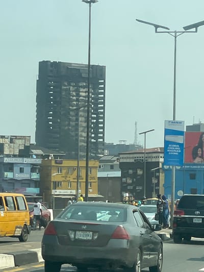 The image captures an urban street scene in Ebute-Metta, Nigeria, under a hazy, light blue sky. A prominent, multi-story skyscraper, heavily damaged and blackened by fire, dominates the upper-left background. Its facade displays extensive charring and visible structural damage, with numerous window openings appearing empty or compromised across its height.
In the midground, below the damaged skyscraper, a cluster of smaller, multi-story buildings lines the street. These include a bright yellow building with multiple windows and "HAIR BEACONS" signage, and a light blue building to its left, displaying "DARLING" branding. Several of these lower buildings feature solar panels on their roofs.
The foreground depicts a multi-lane road with active traffic. Vehicles visible include a yellow commercial minibus (danfo) on the far left, a dark grey Honda sedan (license plate AAA-698MA) in the center, and the front of a black SUV on the right. A red car is partially visible behind the yellow minibus.
Pedestrians are present on the sidewalks and a central median. A male in a white t-shirt and dark trousers stands near the red car on the left. On the right, several individuals, including one wearing a blue garment, are visible on a raised median or sidewalk.
Tall, multi-headed streetlights punctuate the scene. On the right, a large blue advertising billboard features the "DAIKIN" logo and the text "GLOBAL COOLING, NIGERIAN LIVING," alongside a partial image of a woman.