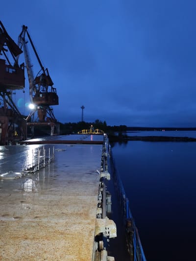 The image presents an industrial port scene at dusk or night, viewed from the deck of a vessel, likely a cargo ship or barge, in Medvezhyegorsk, Russia. The ship's deck in the foreground is wet and appears worn with rust, reflecting artificial lights.

Two large, rusty port cranes dominate the left side of the mid-ground. One crane is brightly illuminated by its own light, and the number "43" is visible on its structure. Reflections from these lights are prominent on the wet deck. Beyond the cranes, a tall, slender tower stands against the dark blue sky, and other smaller, yellow industrial equipment can be seen on the distant dock.

To the right, a dark body of water stretches towards a faint shoreline in the distance, where a few small lights are discernible. The sky above is a deep, rich blue, characteristic of twilight or nighttime conditions. No people are visible in the frame, contributing to a quiet, stark, and industrial atmosphere. Yellow and black striped hazard markings are present on machinery near the ship's railings. The wet surfaces suggest recent rain or a damp environment.