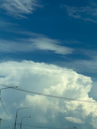 The image shows a wide shot of a sky with large, white cumulonimbus clouds dominating the lower half, against a deep blue sky in the upper half. Several streetlights with dark poles and white fixtures are visible in the foreground, silhouetted against the clouds. Power lines are strung between the poles, also appearing as dark lines against the sky. The scene appears to be an outdoor environment in the city of Zapadny, Russia, likely during daylight hours. No people are visible in the image.