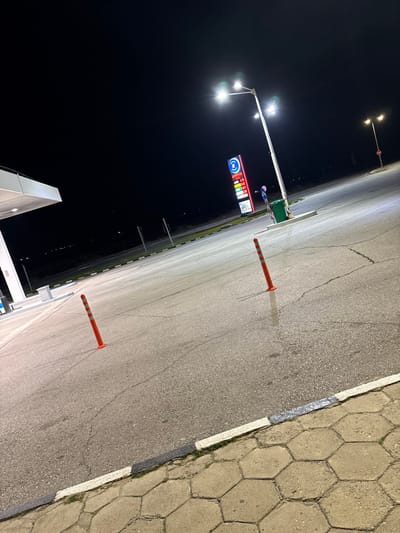 The image depicts a nighttime scene at a gas station in Strumyani, Bulgaria. The environment is dark, illuminated by multiple bright overhead lights mounted on a tall pole. The ground consists of asphalt pavement, showing numerous cracks, with a concrete curb featuring alternating black and white paint separating it from a hexagonal tiled sidewalk in the foreground. Two orange, flexible traffic bollards are positioned upright on the asphalt in the mid-ground.
To the left, a white canopy structure, characteristic of a fuel station, is partially visible. In the background, centrally located, stands a tall, illuminated blue and white sign displaying "BARRELS" at the top, with several fuel prices listed below: 1.99, 2.11, 2.31, and 1.80. Adjacent to the base of the sign and the main light pole is a green trash bin. A single, blurry human figure, dressed in pink or purple clothing, is visible near the base of the fuel price sign, facing away from the camera. The background beyond the gas station shows a dark horizon with faint distant lights. No vehicles are present in the immediate vicinity of the gas station forecourt.
