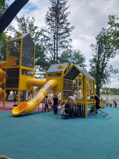 An aerial view of a bright yellow playground with a tubular slide, climbing structures, and a netted enclosure. Several children are playing on the equipment, some climbing, others sliding. The playground is set on a blue and red soft-surface ground, surrounded by trees. In the background, a body of water and a cityscape are visible, hinting at the location in Alikonovka, Russia. The sky is overcast, with scattered clouds.