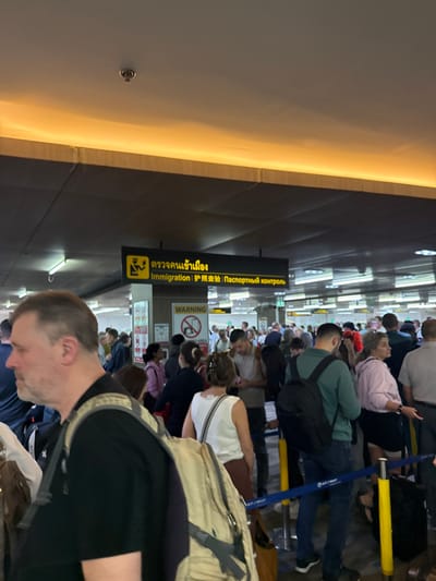A crowded airport immigration line is visible. People are standing close together behind stanchions. Signage in multiple languages (Thai, English, and Russian) indicates "Immigration" and "Passport Control." A warning sign with a "no photo" symbol is also visible. Many travelers are carrying luggage and backpacks. A man is standing in the foreground, wearing a black shirt with tan backpack straps. The lighting in the airport appears bright.