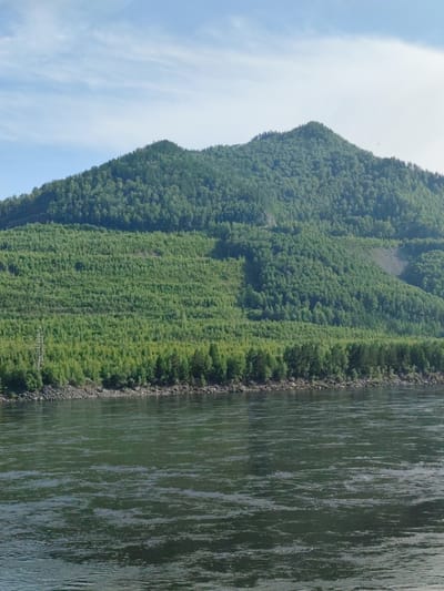 This media file depicts a scenic landscape featuring a wide, dark river in the foreground. The river's surface is rippled, reflecting the sky and surrounding greenery. Along the riverbank, a rocky shore transitions into a dense forest that covers the lower slopes of a large, verdant mountain. The mountain dominates the background, its slopes densely packed with trees. The sky above is a light blue with scattered white clouds. A single, tall electrical pylon is visible on the left side of the image, standing out against the natural backdrop. The scene appears tranquil and evokes a sense of natural beauty. The lighting suggests it is daytime, likely late morning or early afternoon, given the clear sky and bright illumination of the mountain. The location is specified as Cheremushki, Russia, which aligns with the mountainous and forested terrain. There is no visible text within the image.