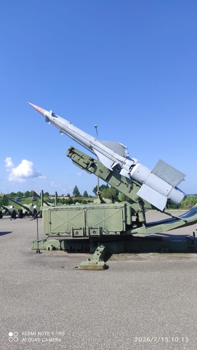 In the city of Лашанскі сельскі Савет, Belarus, a missile launcher with a missile is displayed outdoors on a paved surface. The missile, painted light gray with a red tip, is angled upwards, mounted on a green launcher. The launcher itself is robust, with a large green box-like structure at its base and supporting mechanical arms. In the background, other military equipment, including what appear to be cannons, are lined up, suggesting a military museum or exhibition. The sky is a clear, bright blue with a few scattered white clouds. The overall environment is that of an open, sunny day, typical for an outdoor display.