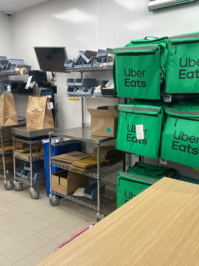 The image displays an indoor commercial staging and storage area, likely within a food service establishment in Chatham, United Kingdom. No individuals are present in the scene.
The area features white tiled flooring and white walls. On the left, a mobile stainless steel rolling cart is visible, topped with a digital scale. On the scale, two brown paper bags are placed, one clearly labeled "Make McDelivery rewarding." The cart's lower shelves contain stacks of brown paper bags and multiple grey plastic storage bins. To the left, a second, partially visible rolling cart is also present. Above these carts, a wall-mounted monitor is visible, along with multiple wall-mounted wire shelves holding numerous grey plastic bins containing various items. A white CCTV camera is mounted in the top-left corner.
In the center, a fixed stainless steel counter is present. Beneath it, additional wire shelving units are stocked with stacks of brown paper bags, flat cardboard packaging, and grey storage bins. An open brown cardboard box rests on one of these shelves. A blue cylindrical bin is positioned under the counter.
The right side of the image is dominated by a vertical array of at least seven bright green Uber Eats insulated delivery bags, each prominently displaying the "Uber Eats" logo. These bags are suspended from a wall-mounted fixture. A white receipt or label is affixed to one of the central Uber Eats bags. A light-colored wooden-patterned countertop occupies the lower right foreground, partially obscuring the base of the delivery bag stack and a portion of the floor.