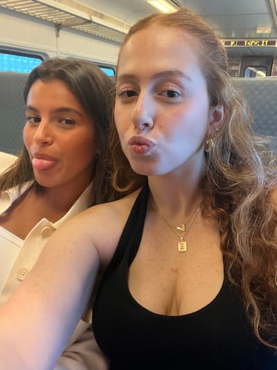 Two young adult females are seated inside an NJ Transit commuter train, likely in or near Linden, New Jersey. The woman on the right, with long wavy reddish-brown hair and a light complexion, wears a black ribbed halter-neck top, two gold necklaces (one with a butterfly pendant, the other with a rectangular "E" initial pendant), and gold hoop earrings. She is looking towards the camera with pursed lips. The woman on the left, with long dark hair and a darker complexion, wears a light-colored button-up jacket over a white top and is looking at the camera with her tongue extended. Both subjects are posing for a casual photograph. The background displays interior elements of a train car, including gray fabric seats, a window, overhead luggage racks, and fluorescent lighting. A partial overhead sign showing "N.J. TRANSIT" is visible on the upper right.