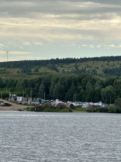 The image depicts a body of water in the foreground, with a shoreline in the middle ground. On the shoreline, a small collection of buildings, a parking area with several vehicles, and a variety of boats covered with tarps are visible. Beyond the shoreline, a dense forest covers rolling hills. In the distance, a tall, lattice-like structure, possibly a radio tower, stands against a cloudy sky. The location appears to be a rural or semi-rural area, possibly a marina or boat storage facility near Krasny Bor, Russia, given the visual cues. The overall scene suggests a quiet, overcast day.