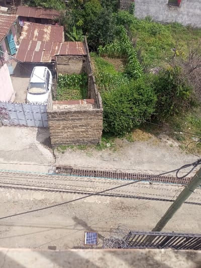 This image, captured from an elevated perspective in Kiamaina ward, Kenya, depicts a detailed urban and semi-rural scene under clear daylight.

The foreground features a cracked concrete and dirt pathway or road. A small, square solar panel rests on the ground towards the bottom-center. To its right, a bundle of barbed wire or razor wire is present. A wooden utility pole with multiple overhead wires extends diagonally from the bottom-right towards the upper-left. Shadows from these wires are cast across the pathway. A metal drainage grate runs horizontally across the mid-foreground, with a light blue hose or cable positioned parallel to it.

In the midground, a substantial wall constructed from light brown stone blocks forms an enclosure or part of a building, topped with a rusty corrugated iron sheet. To the left of this structure, behind a white metal gate, a white SUV or station wagon is parked on a concrete surface. Adjacent to the parked vehicle, within a walled section, a small garden plot contains cultivated green leafy plants. To the right of the stone-block wall, the ground transitions into an area with dense green bushes and scattered vegetation.

The background reveals several structures with severely rusted corrugated iron roofs. Laundry is visible hanging on a line near one of these buildings. Beyond these structures and the immediate garden, a large area of uncultivated or semi-cultivated land extends, featuring various green plants, including tall stalks resembling maize or corn, and several trees. A light-colored building with a dark-framed window is partially visible in the upper-right background, further away. No individuals are visible, and no overt actions or interactions are depicted within the scene.
