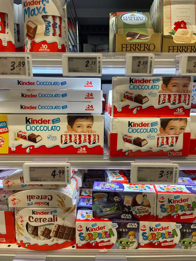 This image depicts a brightly lit confectionery aisle within a retail store in Monsummano Terme, Italy. The scene focuses on multiple shelves fully stocked with various Kinder brand chocolate products, alongside other Ferrero items.

The top shelf on the left displays Kinder Maxi chocolate bars in white and red packaging, indicating "x10" units, priced at 3.89€. Adjacent to this, in the upper right quadrant, are Ferrero Rocher Macadamia Milk chocolates in a gold-brown box, priced at 2.39€, and a white-packaged Ferrero product labeled "Coco & Mandorla."

The middle shelves are predominantly filled with stacks of Kinder Cioccolato boxes. These white boxes feature a child's face peeking over a row of chocolate bars, with "LATTE + CACAO" and "SENZA GLUTINE" labels. Some boxes indicate "x24" units, with one shelf displaying a price of 4.99€ for a Kinder Cioccolato "T24 500G." Other stacks of Kinder Cioccolato are marked "x16" units.

The bottom shelf presents Kinder Cereali chocolate bars in white and brown packaging, showing chocolate bars with cereal grains. To the right, multiple boxes of Kinder Sorpresa (surprise eggs) are visible. These boxes are predominantly white, red, and blue, labeled "SENZA GLUTINE," and some feature "Star Wars" branding with characters like Yoda. A digital price tag above these boxes displays 3.59€ and identifies the product as "FERRERO KINDER CEREAL T/10/200," despite its placement above Kinder Sorpresa.

All product prices are displayed in Euros on electronic white digital tags. The shelves are metallic and well-organized. No individuals are visible in the scene.