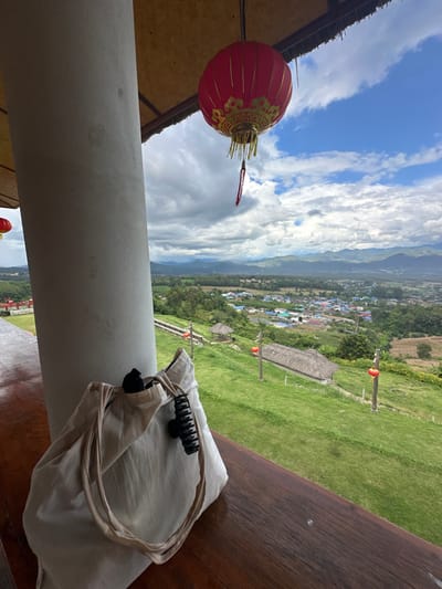 The image displays an elevated vantage point overlooking a valley in Ban Mai Saha Sampan, Thailand. In the immediate foreground, a light-colored cylindrical concrete pillar occupies the left side, beside a dark brown wooden railing or table surface extending horizontally. A light beige fabric tote bag rests on this wooden surface, featuring a black hair claw clip attached to its top edge and a dark object partially visible inside. Suspended from an overhead thatched roof, a prominent red Chinese lantern with gold embellishments and hanging tassels is visible at the top center. A second, partially visible red lantern is located on the far left.
The midground reveals a steep, green grassy hillside descending into the valley. Several evenly spaced wooden posts, each topped with a small red lantern, are positioned along the slope. A small, open-sided gazebo with a thatched roof is visible on the upper part of the hillside, and another building with a thatched roof appears further down the slope. The background showcases a dense cluster of buildings, indicative of a village or small town, situated within the valley, surrounded by agricultural fields and lush vegetation. A chain of mountains and hills defines the distant horizon. The sky above is partially clear blue with significant formations of white and grey cumulus clouds. No individuals are present in the scene.