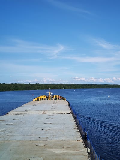 The image shows a large, flat-topped barge or cargo ship moving on a wide, calm body of water. The barge's deck is gray and covered with numerous circular hatches. Along the edge of the deck, a blue railing is visible. In the distance, a dense, dark green tree line lines the horizon. The sky is a clear, bright blue with scattered wispy white clouds. There are no people visible on the deck of the barge. A few small buoys are dotted in the water. The scene appears to be a sunny day, likely in a natural setting with the water body being a river or a lake. There is no information within the image that suggests the location is the city of Shaima, Russia.