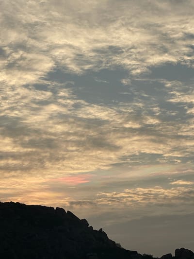 The image captures an outdoor scene in Sanapur, India, at either sunrise or sunset. The sky, occupying approximately two-thirds of the frame, is covered by extensive altocumulus cloud formations, illuminated with warm yellow-orange and subtle pink tones, particularly in the lower left quadrant. Patches of lighter grey-blue sky are visible between the clouds. The lower third of the image features a dark, silhouetted rocky hill range composed of large, irregular rock formations, extending from the left to the right side of the frame. Minimal dark foliage is discernible at the base of the rock formations on the extreme lower right. No individuals, animals, or man-made structures are present.