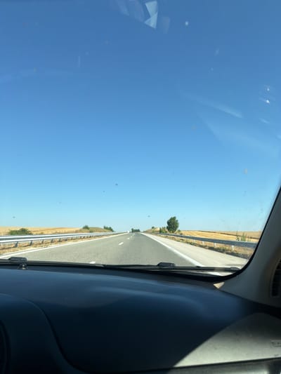 The image is a point-of-view shot from inside a vehicle looking out through the windshield. The vehicle is on a paved highway with a clear blue sky overhead. The highway stretches into the distance, flanked by yellow fields of crops on either side, suggesting a rural landscape. A metal guardrail runs along the side of the road. The dashboard of the vehicle is visible in the foreground, and there are several dust specks or smudges on the windshield. The location is specified as the city of Aleksandrovo, Bulgaria.