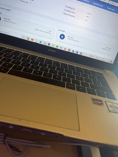 This image captures an angled close-up view of an open HUAWEI laptop computer, situated on a light-colored surface in the city of San José Pinula, Guatemala.
The laptop features a silver-toned body, a black QWERTY keyboard with a Spanish layout, and a touchpad. A red and black AMD Ryzen sticker is affixed to the right palm rest, adjacent to a sticker depicting a wireless connectivity icon.
The laptop screen displays a web page detailing property information. Visible data points include "Median Home Value" at $376,794, a "Median Home Value Rating" of 5/10, a "Family Friendly Score" of 90/100, and a "Walkable Score" of 29/100. Tax billing details show a Zip code of "44216" and "Owner Occupied: Yes." The location "Clinton, OH" is partially visible on the page. Below the main content, a browser bar displays various application icons such as YouTube, WhatsApp, and Spotify. The system tray in the lower right corner of the screen shows the date "27/10/2023," time "14:27," and a temperature of "17°C."
A coiled white cable is partially visible underneath the front edge of the laptop. No individuals or direct interactions are depicted in the scene.
