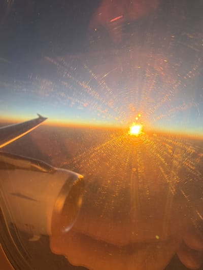 An aerial view from an airplane window shows a vibrant sunset over the city of Talas, Turkey. The sun, a bright, intense orb, is positioned low on the horizon, casting a golden glow across the sky and the clouds below. The light is so strong that it creates a radial pattern of streaks and reflections on the airplane window, resembling concentric circles. The wing of the airplane is visible in the foreground, its metallic surface reflecting the warm hues of the sunset. Part of an airplane engine is also visible, a hulking, light-colored mass. The scene is dominated by warm tones of orange, yellow, and gold, with hints of blue in the upper atmosphere. The ground below is obscured by the atmospheric haze and the intensity of the sunlight, but it appears to be a landscape of urban sprawl.