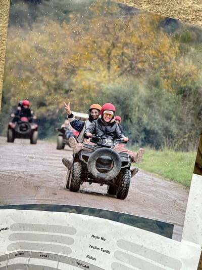 A printed photograph depicts a group of individuals riding all-terrain vehicles (ATVs) on a dirt road in Çiftlikköy, Turkey. In the foreground, two individuals are seated on a black ATV with a spare tire mounted on the rear. The person in the driver's position wears a red helmet, yellow goggles, and a black jacket, smiling and looking towards the viewer. The passenger, wearing an orange helmet, dark sunglasses, and a blue and white jacket, is smiling and making a peace sign with their left hand. Both have their legs extended and feet raised, wearing tan boots. The road surface is wet and muddy, with puddles visible. In the mid-ground, another ATV with two riders, one wearing a red helmet, is visible further down the road. Additional ATVs with riders are faintly visible in the background. The environment consists of dense autumn foliage with yellow and green trees lining both sides of the road. Fine white specks are visible across the image, potentially indicating rain or dust on the photograph itself. The lower portion of the image shows the top edge of a grey document with Turkish text, including "Arşiv No," "Teslim Tarihi," "Kalan," "ADET," "FİYAT," and "TOPLAM."