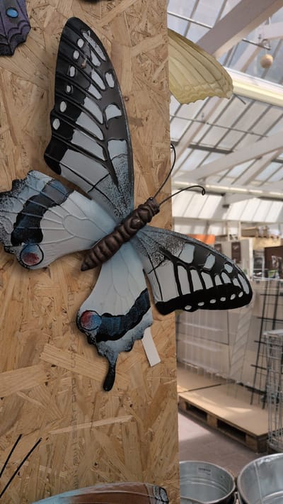 The image shows a decorative butterfly sculpture mounted on a wooden panel. The butterfly has black, white, and blue wings with a segmented brown body. Part of the background shows a greenhouse or store interior with shelves, pallets, and metal bins. The butterfly is the primary subject, and the wooden panel appears to be part of a display. The image was taken in Sömmerda, Germany.