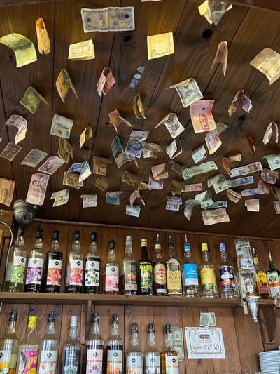 The image depicts a wall adorned with various foreign currencies, possibly a display or a collection of tips left by patrons. The wall itself appears to be made of wood planks. Beside the money-covered wall is a rack of liquor bottles and flavoring syrups. A sign in French, "carte bleue a partir de 2€50," suggests that credit card payments are accepted for purchases starting from 2.50 euros. The specific location is implied to be Montpellier, France.