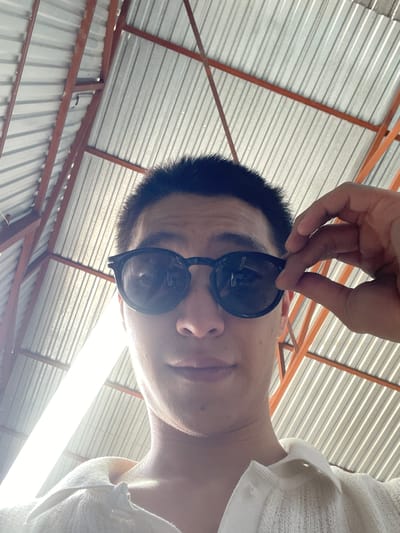 In Кенен Әзірбаев ауылы, Kazakhstan, a low-angle, eye-level shot captures a young person adjusting dark, round sunglasses. The individual is wearing a white collared garment, possibly a sweater or polo shirt. Their dark hair is short. Behind them, a corrugated metal roof is visible, supported by orange metal beams. The scene is lit by a bright light source, likely overhead, which casts highlights on the person's face and clothing.