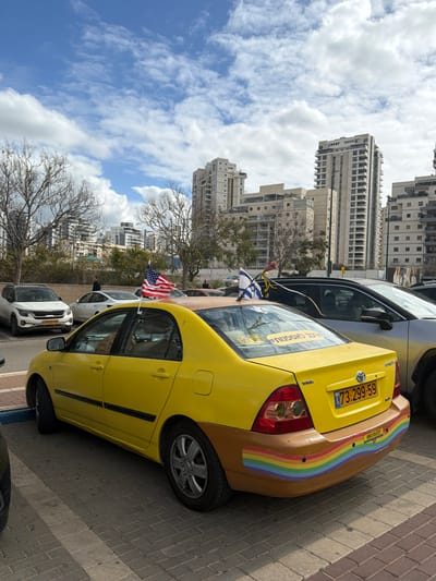 A yellow Toyota Corolla sedan taxi is parked on a paved surface composed of rectangular brick pavers in Kiryat Ono, Israel. The vehicle displays an Israeli license plate "73-299-59" and features a brown rear bumper adorned with a horizontally painted rainbow stripe. A small American flag is attached to the driver's side rear window, while a small Israeli flag is attached to the passenger's side rear window. A yellow sticker with Hebrew text is visible on the rear window. Other vehicles are parked nearby, including a white Kia Seltos and a silver sedan. The background consists of several modern multi-story residential buildings and sparse, leafless trees. A few individuals are visible in the mid-ground, walking or standing. The sky above is blue with scattered white cumulus clouds.