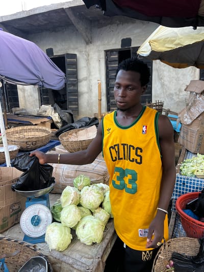 A young Black male stands in an outdoor market in Oko Amakom, Nigeria, wearing a yellow Celtics basketball jersey and black pants. He is holding a black plastic bag filled with items, which he is placing into a metal bowl on a blue scale. In front of him are several heads of cabbage arranged on a table covered with a brown cloth. Behind him, baskets and boxes are visible, along with stalls and buildings that suggest a market environment. Umbrellas, one purple and one yellow, are set up over the stalls. The overall scene depicts a vendor at work in a bustling market.