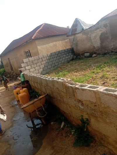 In Gwagwa, Nigeria, a scene unfolds near a structure with a reddish-brown corrugated metal roof. The building's walls are constructed from a tan, textured material. In the foreground, a low wall made of concrete blocks separates a dirt path from a grassy area. Several large, yellow plastic water containers are clustered on the dirt path. A wheelbarrow, also yellow, rests nearby, its contents obscured. Two young boys are present; one in a green shirt stands near the concrete block wall, while the other, in a brown shirt, sits by the water containers, holding a purple plastic cup. A woman walks away from the containers, carrying items over her head, and another person, likely a child, is partially visible behind her. The ground is a mixture of dirt and puddles, suggesting recent rain or water spillage. The overall environment appears to be a residential area under development or in a state of disrepair.