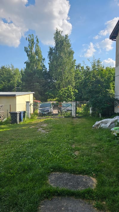 The image depicts a residential backyard in Nemėžis, Lithuania, on a clear day with scattered white clouds. The scene is dominated by lush green grass, a yellow building with a grey roof, and a variety of trees. A dark gray van is visible parked behind a green metal gate. Two trash bins, one blue and one black, are situated near the building. A paved pathway runs through the grassy area. The sun is shining, suggesting it is daytime, likely late morning or afternoon given the bright lighting and clear blue sky. The overall impression is one of a tranquil, suburban outdoor environment.