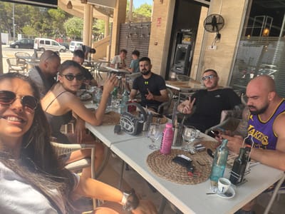 This is a candid, wide-angle shot taken outdoors, likely in Les Meravelles, Spain, given the context. The scene depicts a group of people gathered around a table, enjoying what appears to be a meal or drinks. The overall tone is relaxed and social, with individuals interacting and posing for the camera.

The primary subjects are the people, with several individuals clearly visible. One person on the left wears a purple and gold basketball jersey. Another person in the center wears a black t-shirt with a yellow logo. Several women are also present, one prominently smiling into the camera on the right. The people are of various ages and genders, suggesting a mixed group of friends or family.

The table is laden with various objects, including glasses, bottles (one blue, one dark glass), a straw mat, and what seems to be an ATM machine in the background, indicating a public establishment like a restaurant or cafe. The setting is an outdoor patio or terrace, with wicker chairs and the presence of trees and foliage suggesting a warm, sunny day. The lighting implies it is daytime, possibly late afternoon, with sunlight casting shadows and highlighting some individuals.

The activity appears to be a casual gathering. Some individuals are looking at the camera, while others are engaged in conversation or gesturing. The overall emotion conveyed is one of enjoyment and camaraderie.

Visible text includes "LAKERS" on a jersey, a yellow "ATM" sign, and "CaixaBank" on the ATM machine. The blue bottle has some indistinct writing. The overall image is a snapshot of a pleasant social occasion in a Mediterranean-like setting.