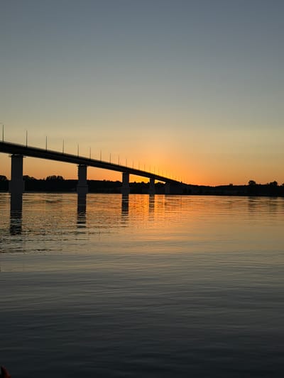 A wide, calm river dominates the foreground of this image, reflecting the warm hues of a setting sun. A long, multi-span bridge stretches across the frame from left to right, supported by numerous tall, cylindrical concrete pillars. The bridge deck is silhouetted against the vibrant orange and yellow gradient of the sunset sky, with streetlights visible along its length. The far bank of the river is lined with a dark silhouette of trees. The scene is set during twilight, likely in the city of Kambarka, Russia, given the context. There are no people or specific actions depicted. The overall impression is one of serene natural beauty combined with man-made infrastructure.
