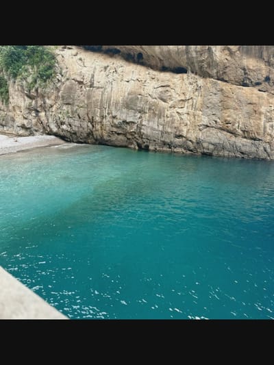A wide shot captures a secluded beach with turquoise waters lapping against a rocky cliff face. The cliff is textured with various shades of brown and grey rock, with sparse green vegetation clinging to its upper reaches. The water is clear near the shore, revealing a pebbly seabed, and deepens to a vibrant blue further out. A small, isolated figure sits on the pebble beach in the upper left quadrant of the image. The foreground shows a sliver of a concrete structure, likely a pier or seawall. The scene evokes a sense of natural beauty and tranquility, suggesting a remote coastal location. This image is not located in Norcia, Italy, as Norcia is a landlocked town in Umbria, not a coastal city.
