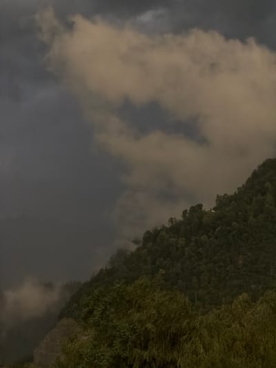 The image depicts a mountain landscape with a dense forest covering the slopes. The sky is filled with dramatic, dark clouds, suggesting an impending storm or overcast weather. Patches of lighter, possibly sunlit, clouds are visible, creating a contrast with the darker areas. The overall atmosphere is moody and perhaps a bit ominous. The location in the city of Cluses, France, indicates this is a view of the surrounding mountainous terrain. There are no people, specific objects, or actions identifiable in the image, making it a purely environmental and atmospheric scene.