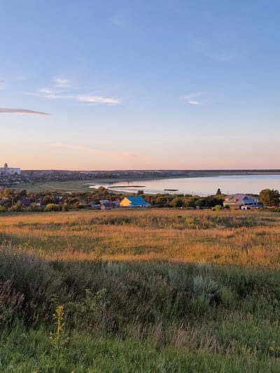 The image depicts a wide, landscape view of a rural village situated along a body of water, likely a river or lake, in Kashgar City, China. The scene is bathed in the warm, soft light of either sunrise or sunset, indicated by the golden hues reflecting off the tall grasses and the pastel colors in the sky. The sky is a pale blue with wisps of clouds, suggesting fair weather.
The village is a collection of houses with various roof colors, including a prominent bright blue roof on one structure. These homes are nestled amongst trees and appear to be situated on a slight incline leading down to the water. The foreground is dominated by a field of tall, dry grass, illuminated by the angled sunlight, creating a textured, golden-brown appearance. Further back, closer to the village, the grass is greener and denser, with some areas appearing more like scrubland. The water stretches across the middle ground, calm and reflecting the sky.
The overall impression is one of tranquility and rural life. There are no people or overt activities visible, contributing to a peaceful atmosphere. The composition, with the sky on the left and the landscape on the right, is unusual due to the image being rotated. The location context of Kashgar City, China, places this scene within a specific geographical and cultural setting, though there are no explicit visual cues to identify it as such beyond the provided metadata. There is no visible text in the image.
