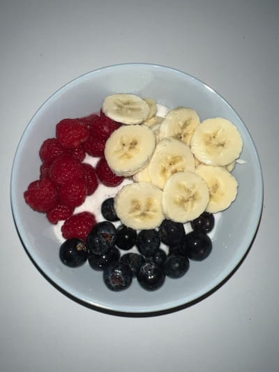 A light blue bowl contains a serving of yogurt topped with fresh fruit. Sliced bananas line the upper-left portion of the bowl. Bright red raspberries are clustered at the bottom-left, while dark blueberries fill the right side. The yogurt is visible between the fruits, providing a creamy base. The bowl is sitting on a white surface, which creates a clean backdrop.