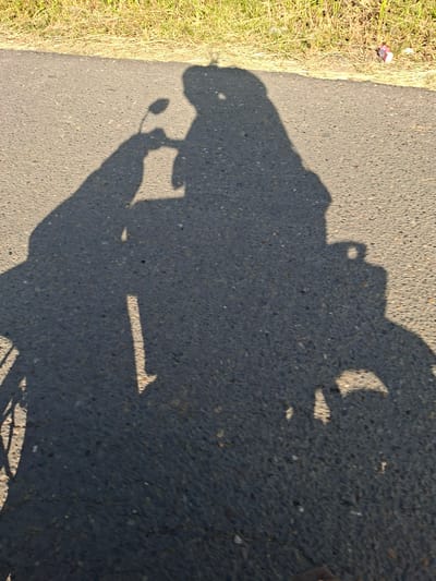 The image shows the shadow of a person riding a motorcycle on a paved road next to a grassy verge. The location is inferred to be in the city of Uleeblang, Indonesia, but is not explicitly verifiable within the image. There is an object on the grassy verge, but it is unidentifiable. The road appears to be asphalt.