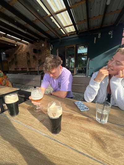 The scene depicts two individuals seated at a wooden slatted table on an outdoor patio in Hornsea, United Kingdom. Overhead, a translucent corrugated roof supported by wooden beams casts distinct striped shadows onto the table and floor.
A young male with light brown, curly hair and a short-sided haircut, wearing a light purple t-shirt, is positioned on the left. He leans forward, looking down at the table, with his right arm resting on the surface. His left hand holds a stemmed glass containing an amber-colored beer.
To his right, a young female with blonde hair, wearing a light grey zipped hoodie, sits with her right hand near her mouth/chin. She is smiling or laughing, with her eyes partially closed. Her left arm rests on the table.
On the table in the foreground are multiple beverages. From left to right, partially obscured, is a glass of dark stout beer with a foam head. Centrally, a tall glass of dark stout labeled "Guinness" also features a foam head. The stemmed glass held by the male contains an amber liquid. To the right, near the female, is a clear glass of water containing ice and a dark straw. Between the two individuals, a light blue or white smartphone with a patterned case (depicting repeating white figures) rests flat on the table.
The background consists of dark, horizontally slatted benches behind the individuals. Beyond the benches on the left is a reddish-brown brick wall adorned with several clusters of hanging green plants. On the right, a solid dark teal wall features a set of double glass doors that reflect outdoor greenery. Natural light illuminates the scene, creating strong contrasts and shadows.