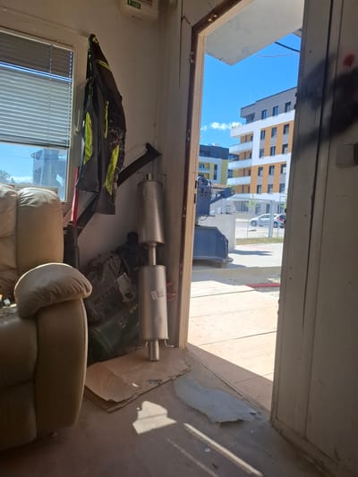 The image shows the interior of a room looking out through an open doorway. The room appears to be under construction or renovation, with exposed materials and debris on the floor. In the foreground, there is a section of a light-colored recliner chair. To the left, a window with horizontal blinds is visible, and a dark jacket with bright yellow accents is hanging on the wall. Two silver exhaust mufflers are stacked vertically near the center of the room, resting on cardboard. Behind the mufflers, what appears to be part of an engine or machinery is visible.

Through the doorway, a bright, sunny outdoor scene is depicted. Several multi-story apartment buildings with modern architecture, featuring white, grey, and orange facades, are visible. A light-colored sedan is parked on the street in front of a chain-link fence. The ground outside is paved with concrete and there is some red-colored tubing visible on the ground. The sky is clear and blue.

The location is identified as České Budějovice, Czechia. The scene suggests a residential construction site, possibly an apartment complex, viewed from a temporary on-site room or storage unit. There are no people visible in the image.