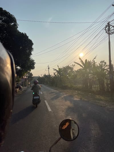 This image captures a street scene in Kebonan, Indonesia, likely during sunset given the low angle of the sun. The perspective is from the back of a motorcycle, with the rider's helmet visible in the foreground on the left. Ahead, another motorcycle is traveling away from the camera, ridden by a person wearing a light-colored jacket and a pink helmet. The motorcycle's license plate is visible, reading "AD 6880 CH". The road is asphalt with a white lane marker. On the right side of the road, there are numerous power lines strung between poles, with a utility pole prominently featured. Lush vegetation, including banana trees, lines the right side of the road, with some buildings and trees visible on the left. The sky is clear with a warm, golden hue, indicating the sun is low on the horizon. The overall atmosphere is peaceful and rural.