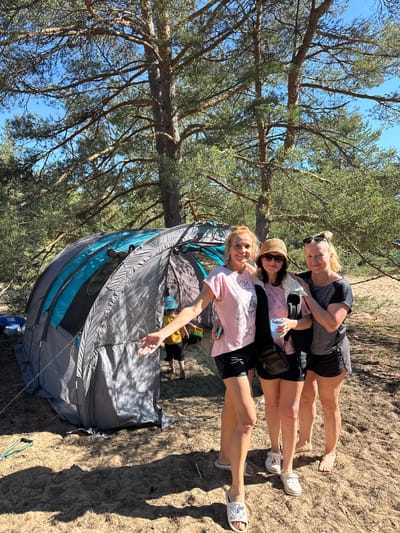 Three women stand in front of a grey and blue tent on a sandy area surrounded by trees. The woman on the left wears a pink t-shirt and black shorts, her left arm extended towards the camera. The middle woman wears a pink t-shirt, black jacket, and a straw hat, holding a blue cup. The woman on the right wears a black t-shirt and black shorts, with sunglasses on her head. A young child in a blue hat and yellow shirt is partially visible inside the tent. The environment appears to be a campsite in a wooded area with sandy ground. The sky is blue and sunny.
