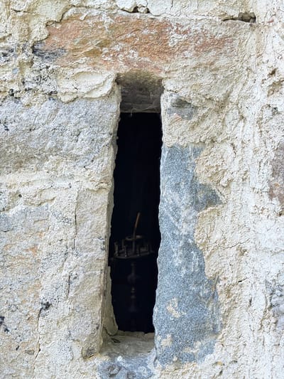 A stone wall in Govedartsi, Bulgaria, contains a rectangular opening. Within the opening, a dark space is visible, with a metal gear and rod partially visible. The stone wall is textured, featuring variations in color from gray to white with some hints of brown. Cracks and crevices are evident on the wall's surface.